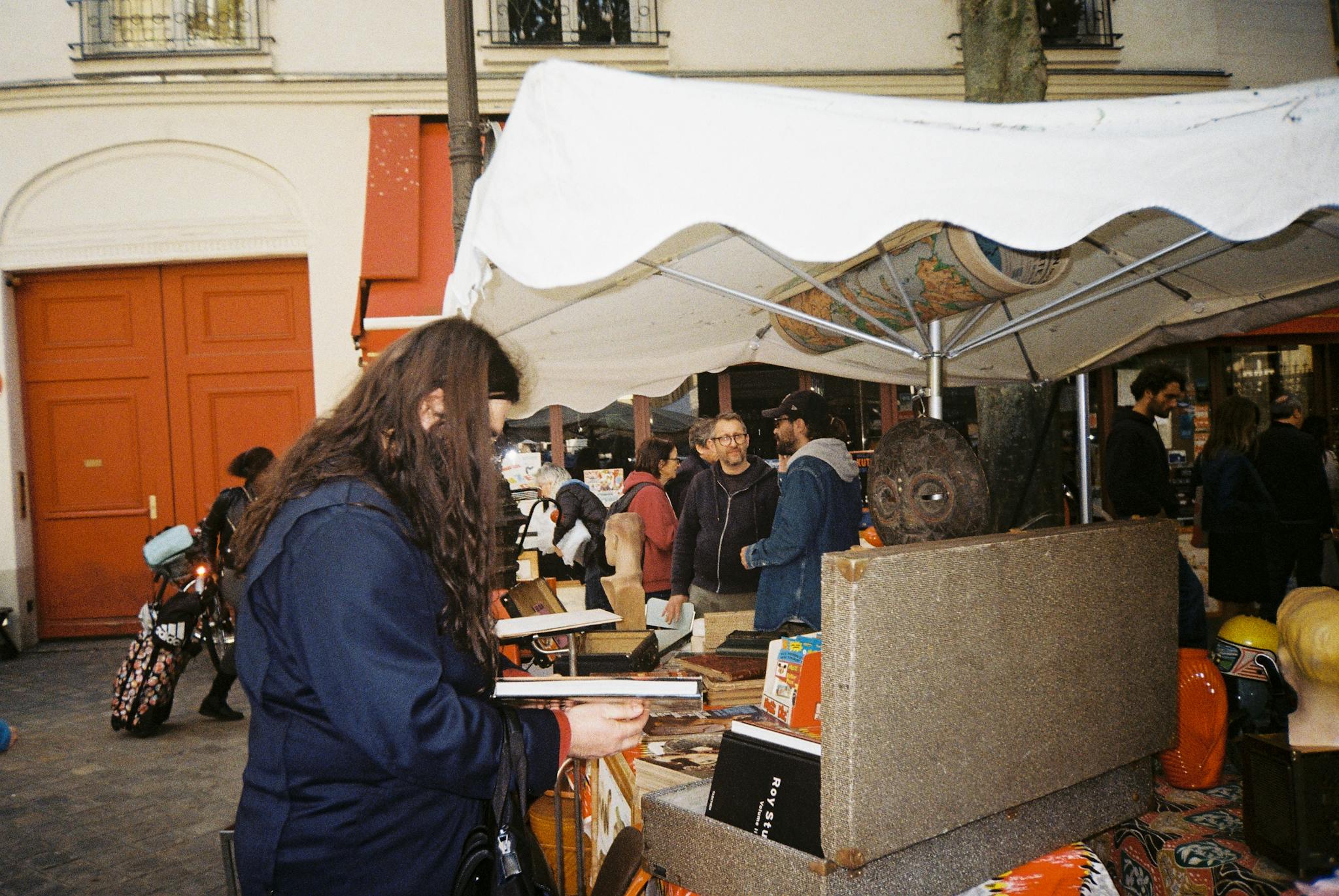 A lively street market scene showing diverse people shopping at outdoor stalls in an urban setting.