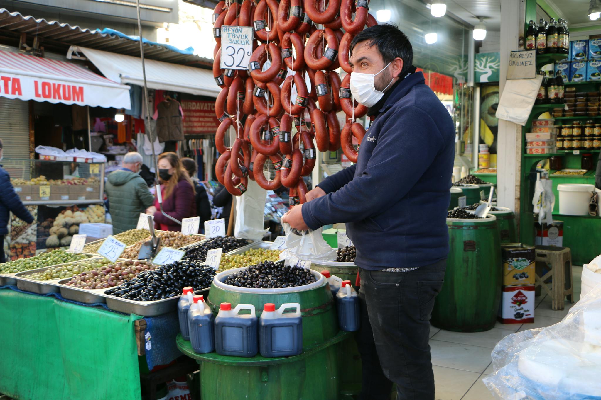 Vendor wearing a face mask selling olives and sausages at a bustling street market.